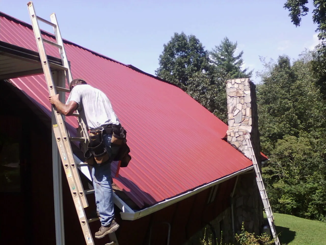 Roofer on a ladder beside a red metal roof and stone chimney — Soffit & Fascia Repair by Roofing Brothers in Cohoes